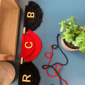Handmade macramé wall hanging with RCB letters on red and black semicircle designs, mounted on a wooden rod