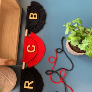 Handmade macramé wall hanging with RCB letters on red and black semicircle designs, mounted on a wooden rod