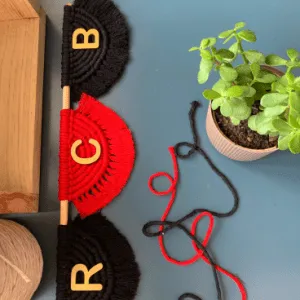 Handmade macramé wall hanging with RCB letters on red and black semicircle designs, mounted on a wooden rod