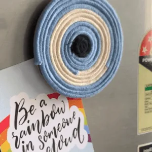 Handmade circular macramé evil eye fridge magnet in shades of blue, off white, and black, displayed on a refrigerator